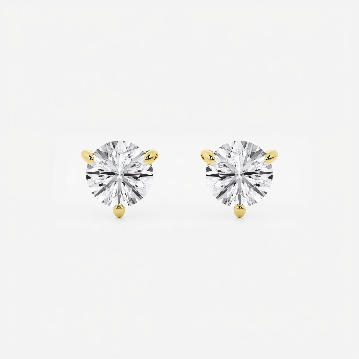 The image shows two stud earrings made from yellow gold. Each earring features a single round brilliant-cut diamond held in a three-prong setting that tapers elegantly toward the stone. Designed to be worn on the earlobes, the studs are shown unattached and arranged side by side on a seamless white surface. Crisp, even studio lighting creates a high-key look with soft shadows beneath the earrings, emphasizing their sparkle and symmetry.  | Adorn By You