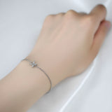 A single delicate silver chain bracelet features a tiny star-shaped charm set with clear marquise and round stones at its center. It\u2019s worn loosely on a slender wrist, with the hand resting gently on a smooth white silk surface. The lighting is soft and diffused, casting an even glow that enhances the polished metal and sparkling facets without harsh shadows. Shot in close-up with a shallow depth of field, the image has a minimalist, elegant product-photography style. | Adorn by You