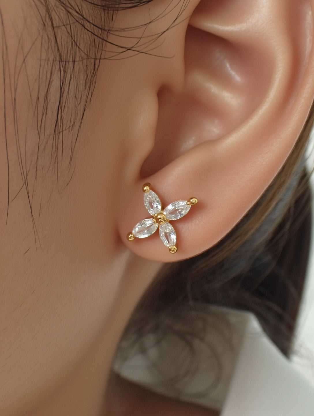 A single gold stud earring featuring four marquise-cut clear gemstones arranged in a cross shape is set in yellow gold. It is worn on the model left earlobe and displayed in a tight profile headshot with dark hair pulled back. Soft, even lighting accentuates the sparkle of the stones against the wearer on smooth skin and white collar. The neutral background and crisp focus give the image a minimalist, editorial feel. | Adorn by You