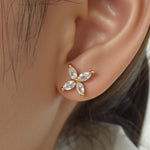 A single gold stud earring featuring four marquise-cut clear gemstones arranged in a cross shape is set in yellow gold. It is worn on the model left earlobe and displayed in a tight profile headshot with dark hair pulled back. Soft, even lighting accentuates the sparkle of the stones against the wearer on smooth skin and white collar. The neutral background and crisp focus give the image a minimalist, editorial feel. | Adorn by You