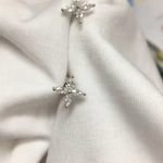 Two silver stud earrings with four clear, marquise-cut stones set in a prong-style cross are nestled into a crisp white fabric. The gemstones sparkle softly under diffused, natural light, their facets catching gentle highlights. Captured in a close-up with a shallow depth of field, the earrings are displayed pinned to the folds of the cloth against a softly blurred indoor background. | Adorn By You