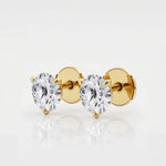 The image shows two stud earrings made from yellow gold. Each earring features a single round brilliant-cut diamond held in a three-prong setting that tapers elegantly toward the stone. Designed to be worn on the earlobes, the studs are shown unattached and arranged side by side on a seamless white surface. Crisp, even studio lighting creates a high-key look with soft shadows beneath the earrings, emphasizing their sparkle and symmetry. | Adorn By You