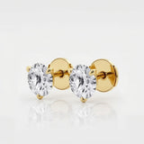 The image shows two stud earrings made from yellow gold. Each earring features a single round brilliant-cut diamond held in a three-prong setting that tapers elegantly toward the stone. Designed to be worn on the earlobes, the studs are shown unattached and arranged side by side on a seamless white surface. Crisp, even studio lighting creates a high-key look with soft shadows beneath the earrings, emphasizing their sparkle and symmetry. | Adorn By You